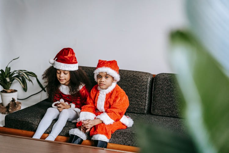 Sincere Black Children In Santa Costumes Resting On Sofa