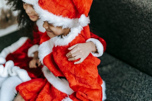 Crop cheerful African American kids wearing red Santa costumes and hats embracing and sitting on cozy couch