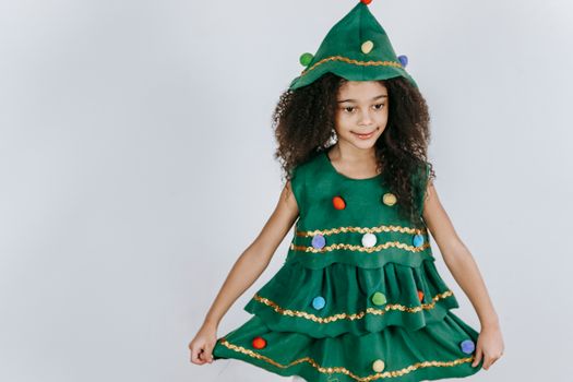 Cheerful African American girl wearing creative Christmas tree costume and hat touching skirt and looking away in light studio