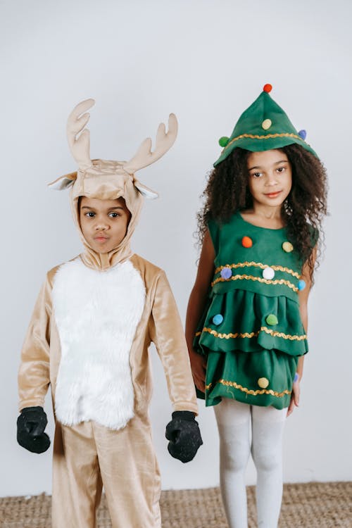 Cheerful African American boy in funny deer costume standing near girl in green Christmas tree dress in light studio