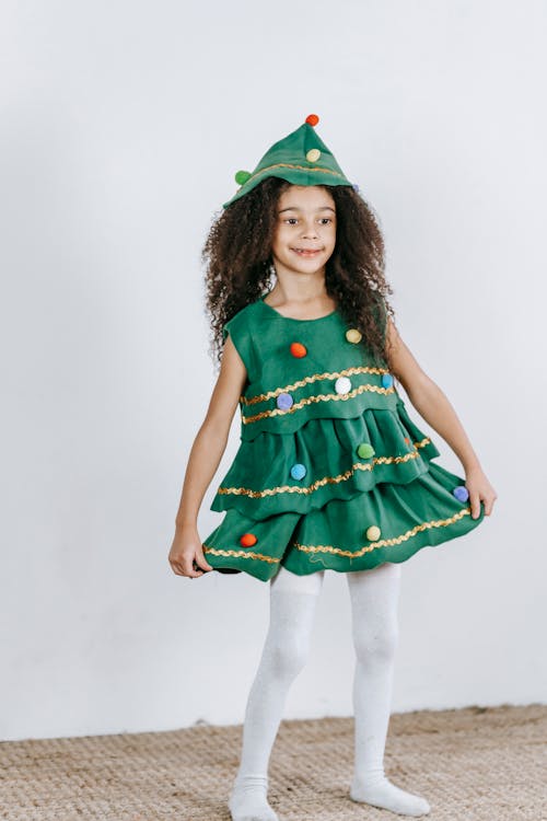 Funny African American girl in creative Christmas tree costume and hat touching skirt and looking away happily while standing against white wall
