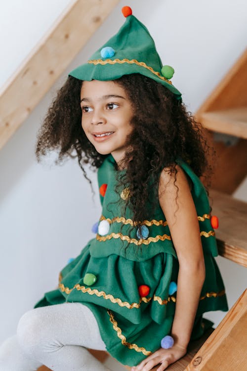 Free Cheerful African American girl with black hair wearing festive green Christmas tree dress and headdress sitting on wooden staircase during holiday celebration Stock Photo
