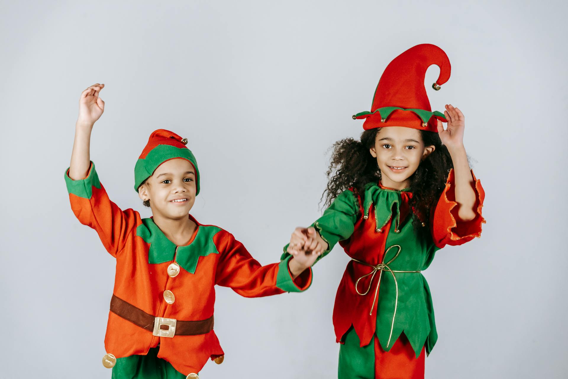 Optimistic African American siblings wearing colorful elf outfits with hats holding hands while standing on white background during Christmas holiday