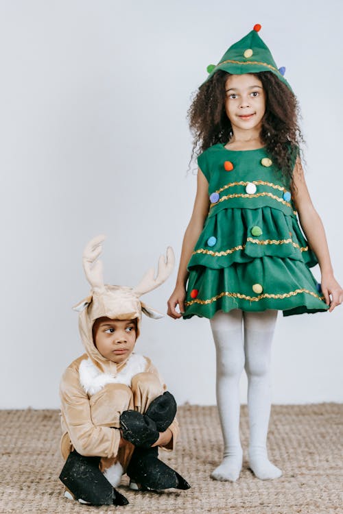 Free Full body of adorable African American girl in Christmas tree dress looking at camera while standing near black boy in reindeer outfit Stock Photo