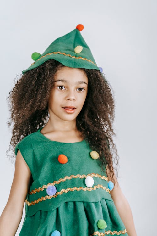 Adorable African American girl wearing headdress and green festive dress looking away while standing on white background during Christmas holiday