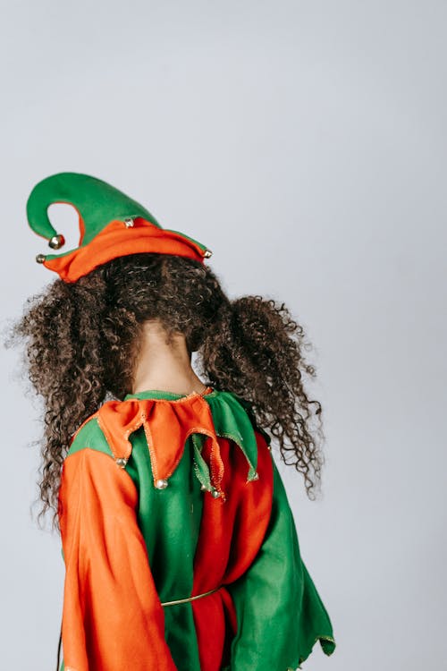 Free Back view of unrecognizable girl with curly hair wearing colorful elf outfit with headdress standing on white background during Christmas holiday Stock Photo