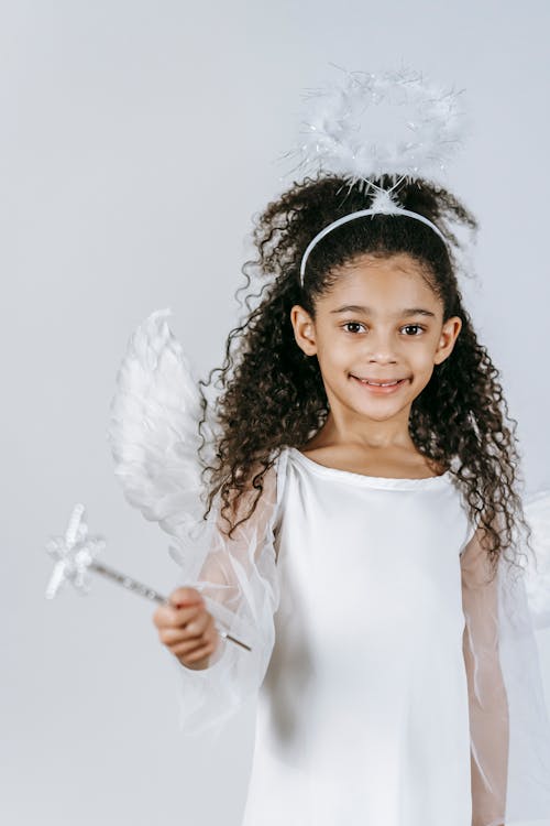 Adorable little African American girl in costume of angel smiling and looking at camera on white background