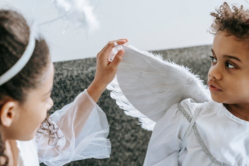 Crop anonymous African American girl playing with wing of angel costume of little cute brother looking away