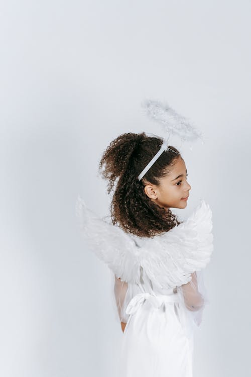 Back view of adorable ethnic little girl with long curly hair demonstrating white wings while standing in studio in angel costume