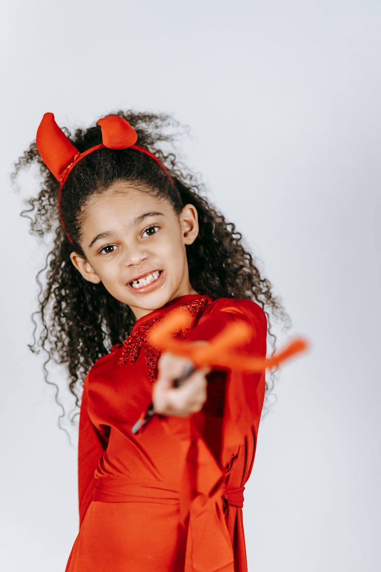 Cute African American Child In Devil Costume And Horns Showing Pitchfork In White Studio