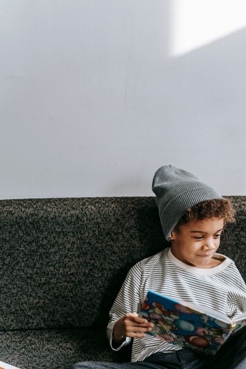 Free Happy little African American boy in casual clothes and hat sitting on sofa and reading book in daylight Stock Photo