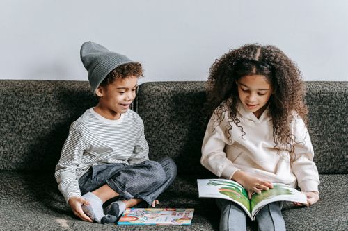 Free Adorable African American girl with little brother in stylish clothes sitting together on sofa and looking at pictures in book Stock Photo