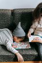 Adorable little black boy in casual clothes and hat lying on couch near crop unrecognizable sister reading fairytale at home