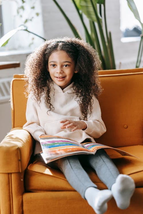 Cute little black girl resting on sofa with book in sunlight