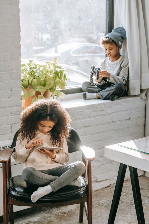 Free Full body of calm African American girl sitting on armchair and browsing tablet during weekend at home with little brother playing with toy on windowsill Stock Photo