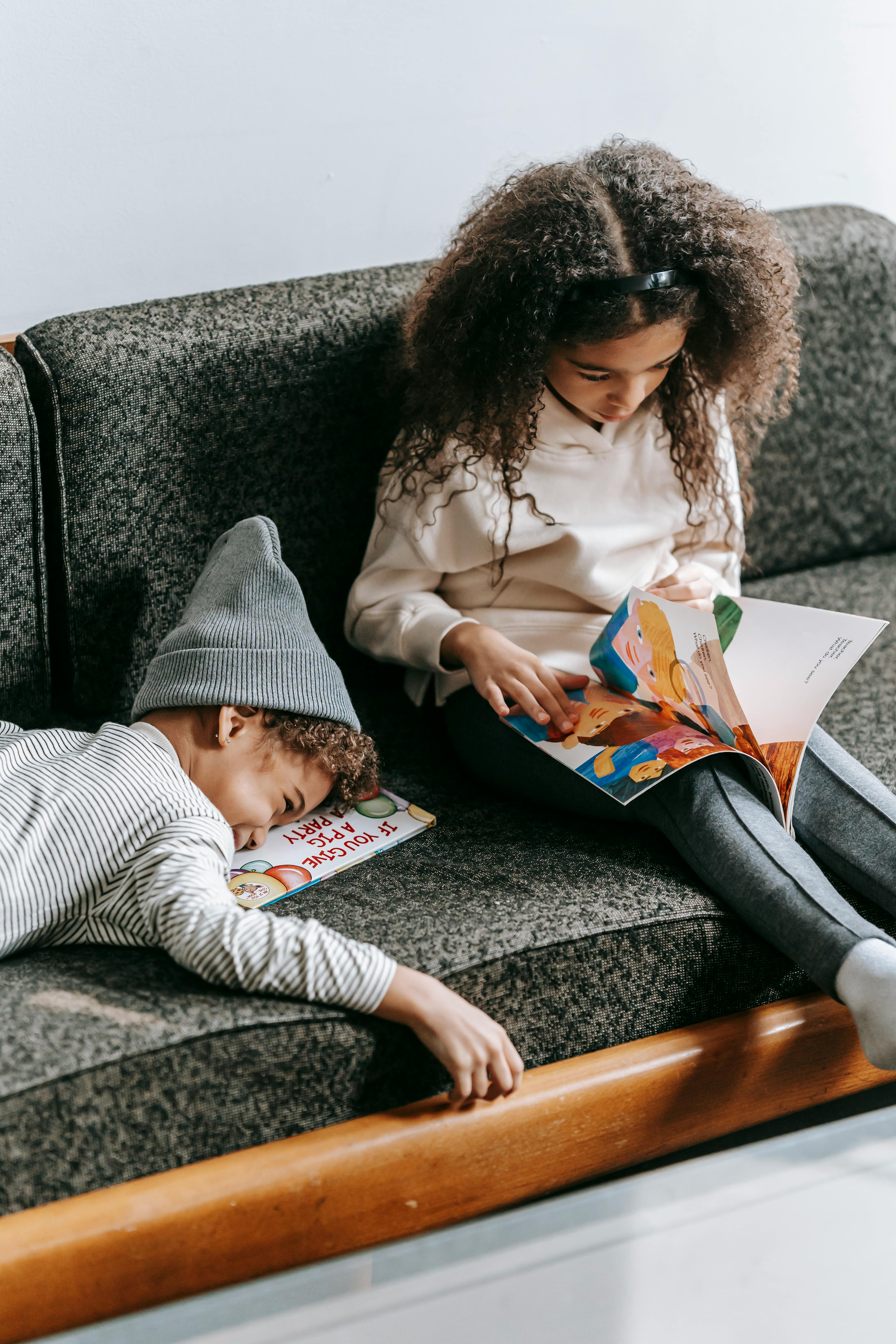 serious ethnic girl reading fairytale near joyful little brother lying on couch