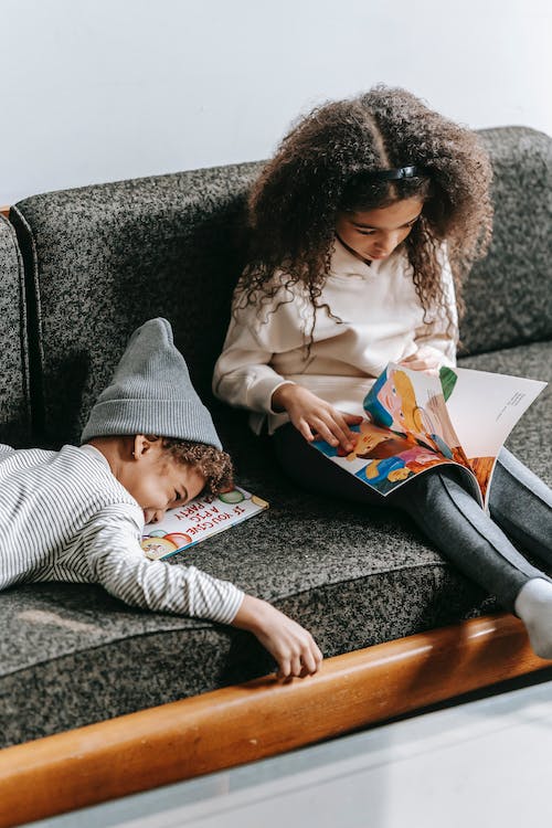 Free Serious ethnic girl reading fairytale near joyful little brother lying on couch Stock Photo