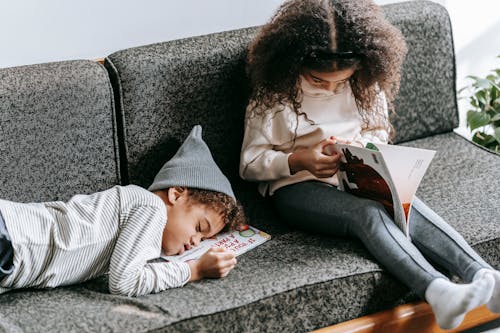 Free High angle of cute African American kid in casual clothes sleeping on sofa near focused sister studying interesting book during weekend at home Stock Photo