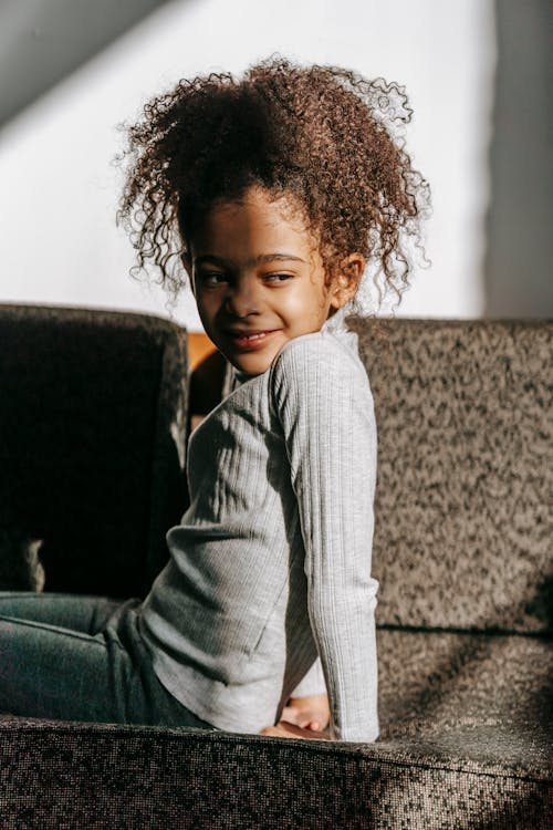 Side view of content little African American child with dark curly hair in casual clothes relaxing on comfortable sofa and looking away