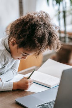 Side view of thoughtful African American schoolgirl with curly hair writing in notebook while sitting at table with laptop and doing homework assignment with concentration