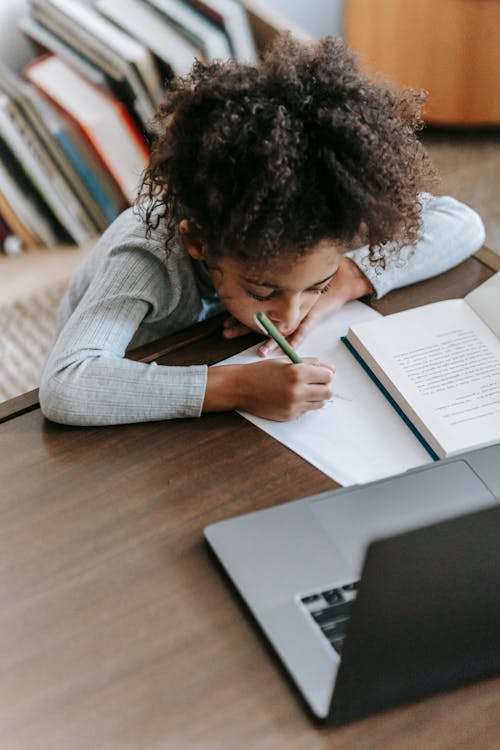 Attentive little ethnic girl writing and browsing laptop while doing homework