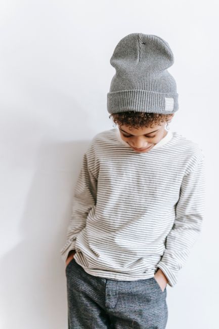 Calm African American boy in trendy outfit standing with hands in pockets and looking down against white wall in room