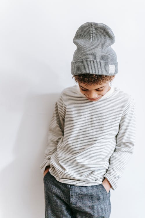 Free Calm African American boy in trendy outfit standing with hands in pockets and looking down against white wall in room Stock Photo