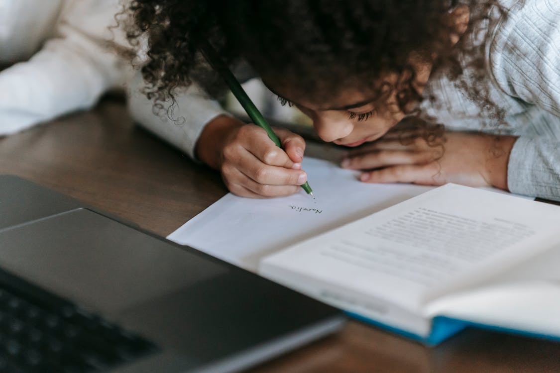 Free Black girl writing on paper while reading book Stock Photo