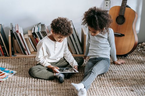 Free Thoughtful African American children in casual clothes sitting on floor against guitar and books and using tablet at home in daytime Stock Photo