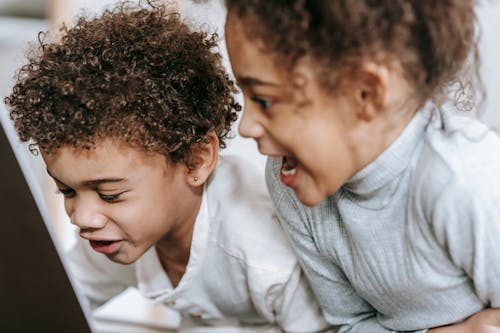 Side view of happy African American siblings watching funny video on netbook while spending time in light room on blurred background