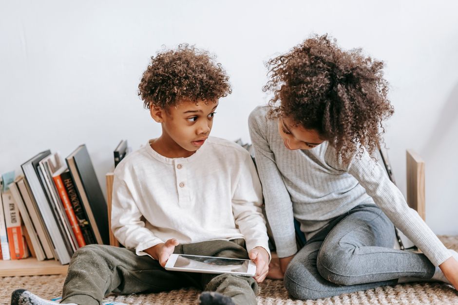 Two children using a tablet indoors, surrounded by books, engaging in digital learning.