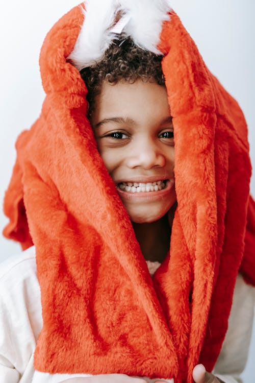 Happy African American boy with red Santa Claus costume on head smiling and looking at camera while standing on white background during Christmas celebration