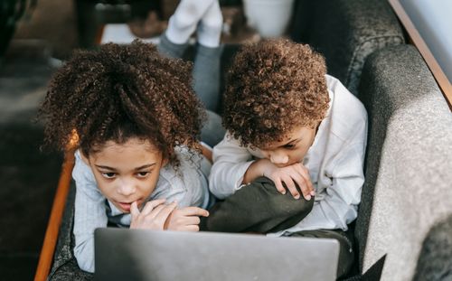 Free Concentrated African American siblings looking at screen of netbook while lying on couch and watching interesting video in light living room Stock Photo