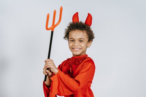 Happy African American boy in red bright costume of devil with pitchfork smiling and looking at camera on white background