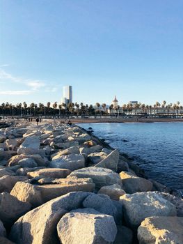 A picturesque view of Barcelona's waterfront with rocky shoreline, palm trees, and city skyline.