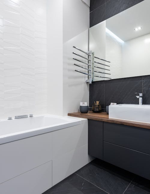 Interior of modern washroom with mirror near white ceramic bath placed near black cupboard with sink and toiletry at home