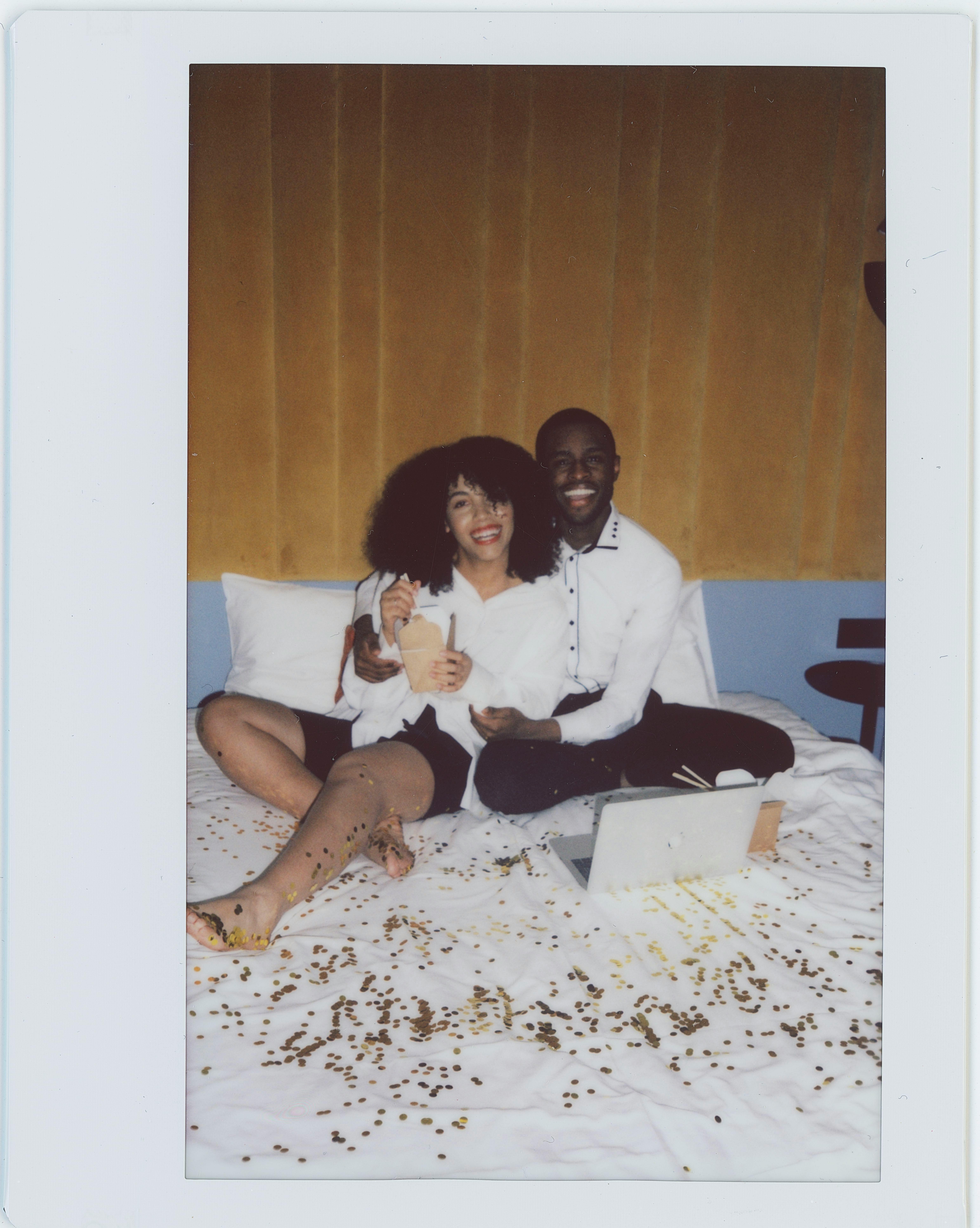 Smiling couple sharing takeout on a bed surrounded by confetti, creating a joyful moment.