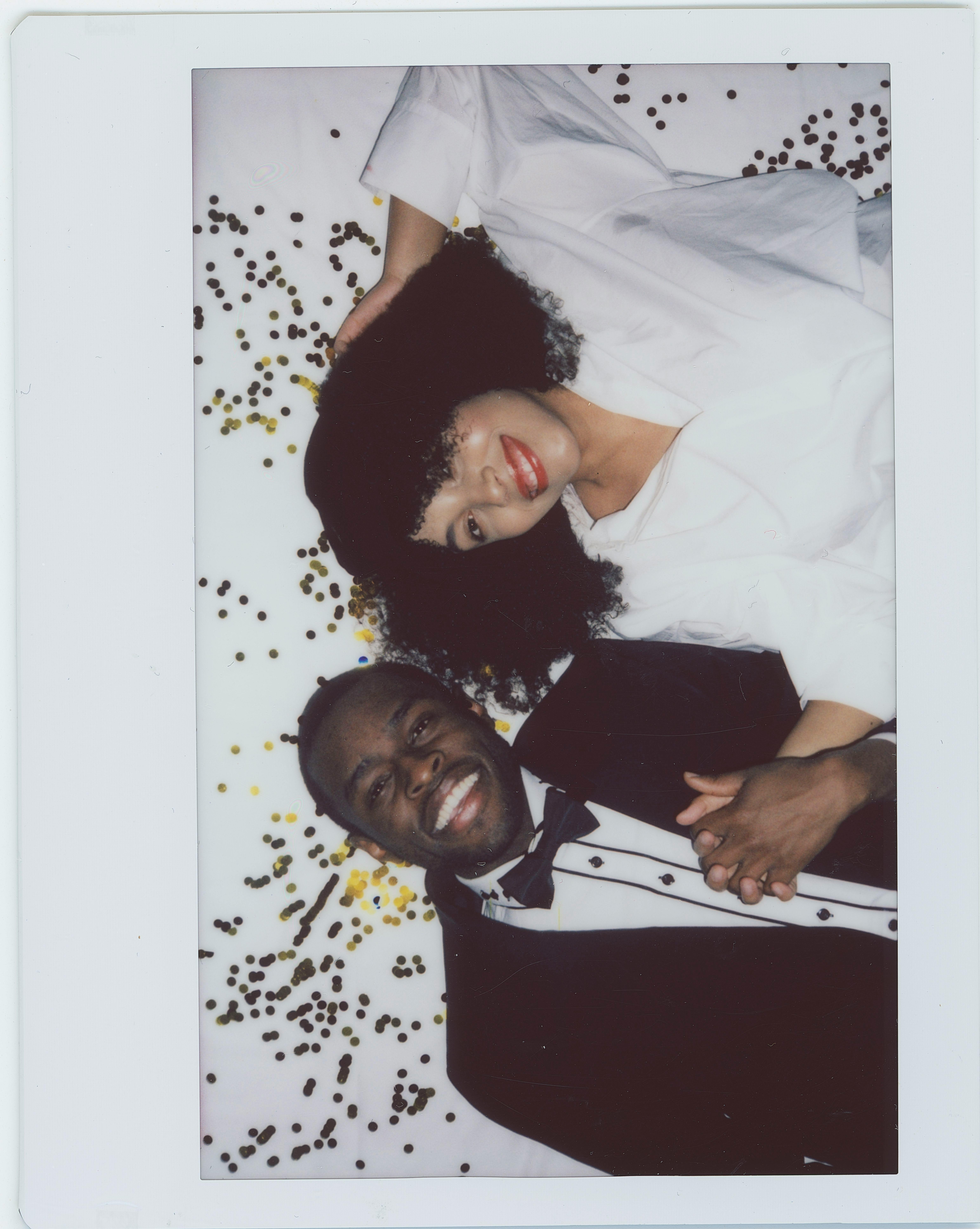 A cheerful couple lying on confetti with formal attire, holding hands and smiling.
