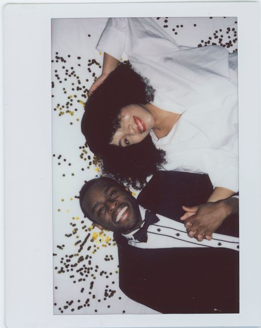 A cheerful couple lying on confetti with formal attire, holding hands and smiling.