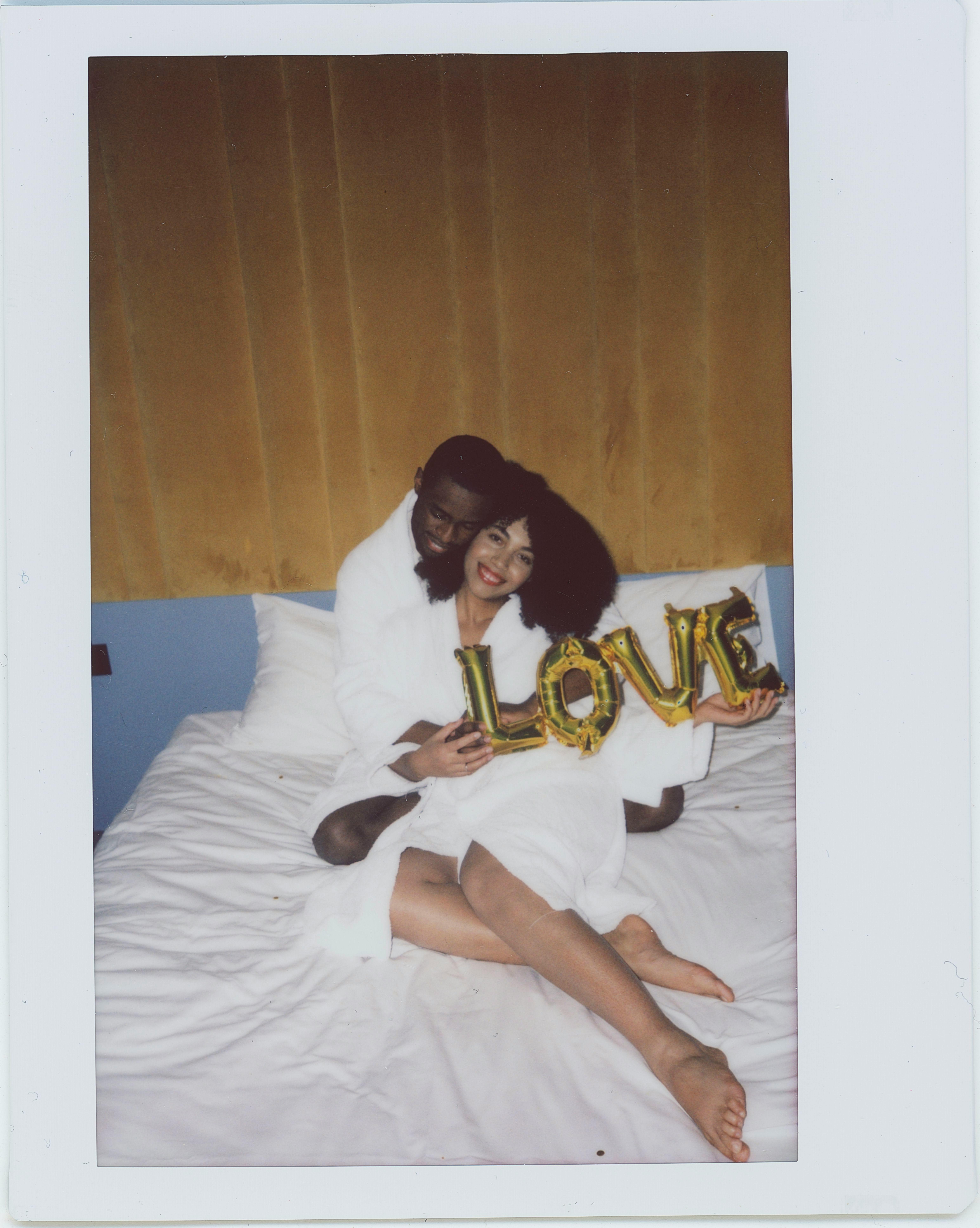 Happy couple in bathrobes sitting on bed, holding love balloon. Embracing each other in cozy bedroom setting.