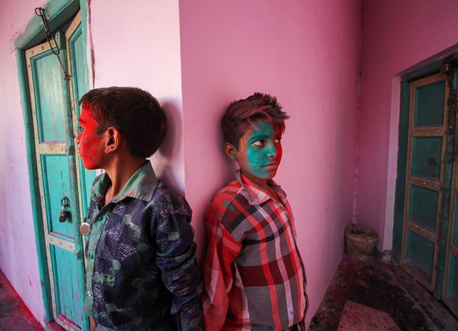 Colorful celebration of Holi Festival with two boys covered in vibrant face paint leaning against a pink wall.
