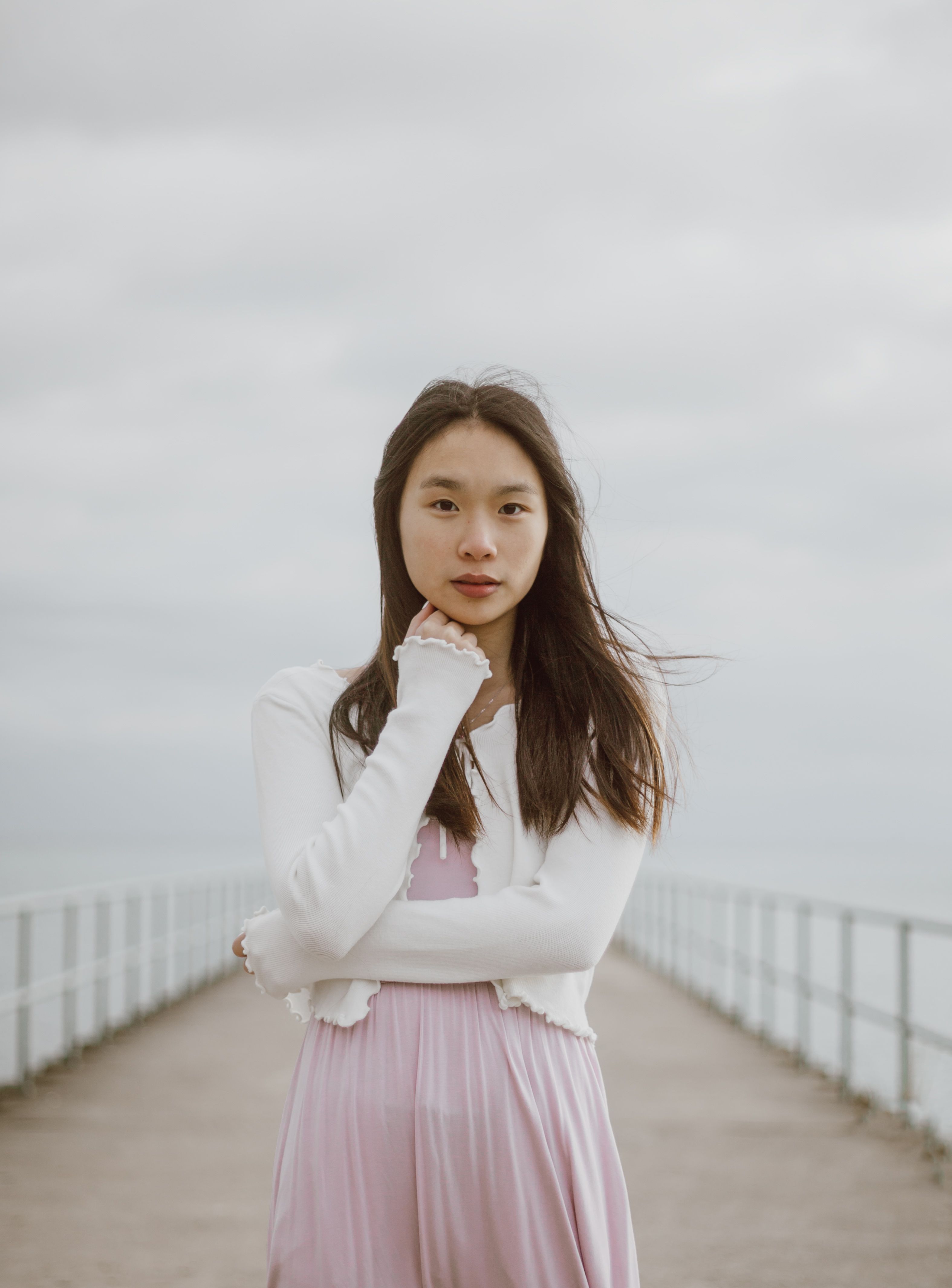 Young tender ethnic female tourist in casual clothes touching chin and looking at camera on fenced embankment between sea