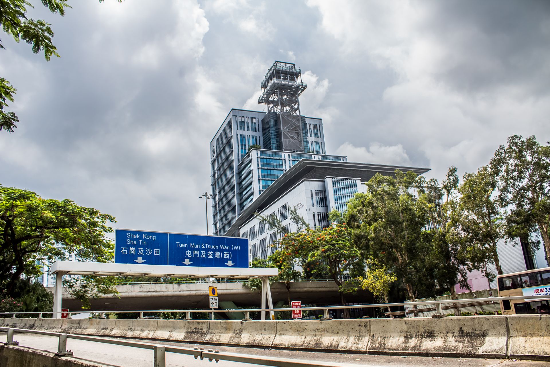 A modern building in Hong Kong on a cloudy day with directional signs and trees in the foreground.