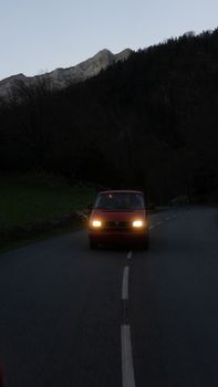 Aged red car with glowing headlights driving on asphalt road near lush green forest growing on mountain slope under cloudless evening sky