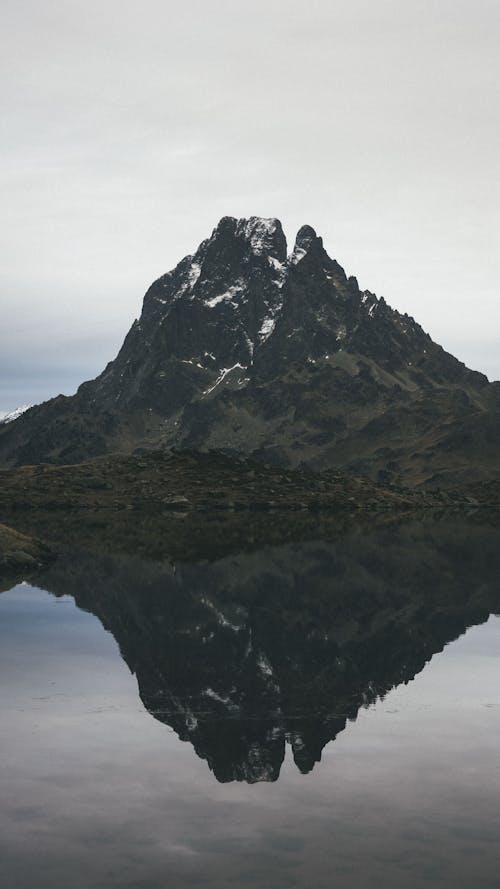 Picturesque landscape of rocky mountain with snowy peak located on shore of calm lake against cloudy sky