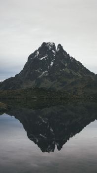 Picturesque landscape of rocky mountain with snowy peak located on shore of calm lake against cloudy sky