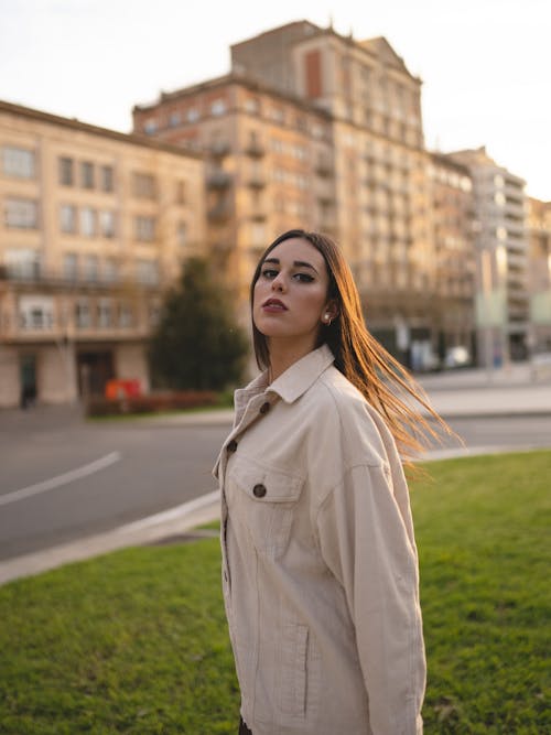 Side view of self assured young lady with long dark hair in trendy outfit standing at roadside near modern building and looking at camera