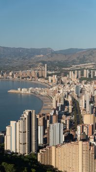 From above of various multistory residential buildings located near sandy beach of sea surrounded by hills under cloudless blue sky in Benidorm