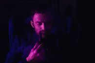 Thoughtful young bearded male in elegant suit smoking pipe while resting in armchair in dark room with purple neon lights