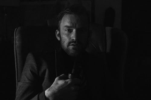 Black and white of serious young male bearded man in stylish elegant suit smoking pipe while resting in comfortable armchair and looking at camera